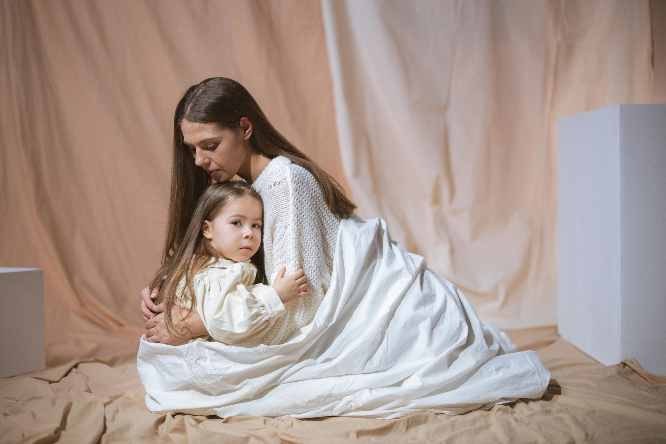 A loving mother and child embrace in a cozy indoor setup, wrapped in a white blanket.